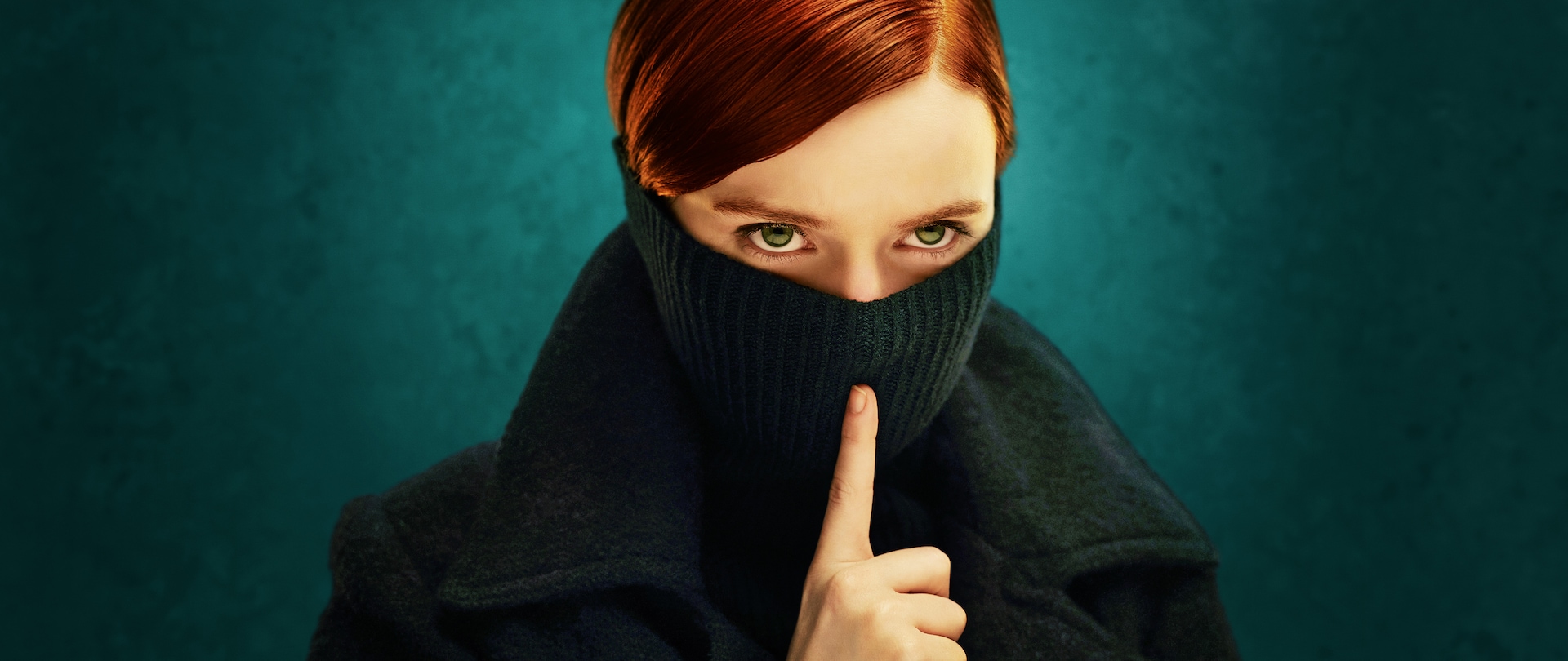 Woman with red hair and green eyes wearing a black turtleneck over her face and shushing.