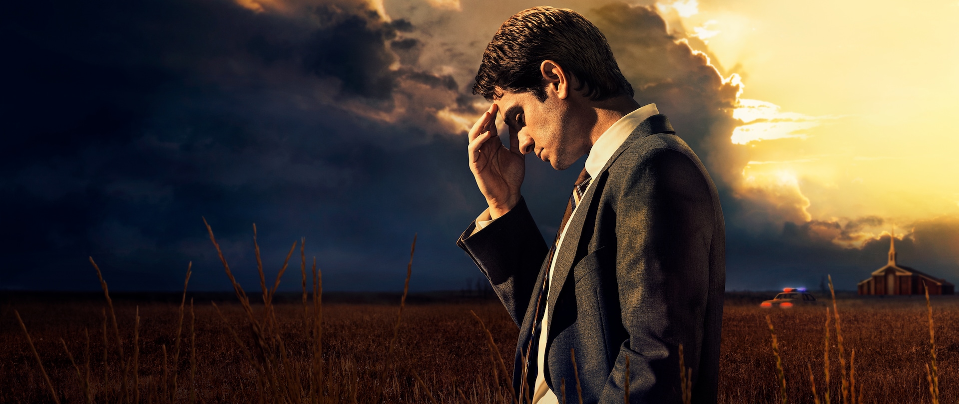 Andrew Garfield stands in middle of a field at the forefront with a church building and police car in the background