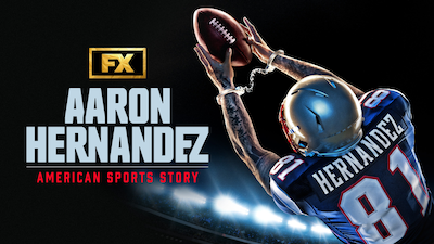 Aaron Hernandez in blue and white football uniform catching a ball with hands in silver handcuffs in a stadium at night.