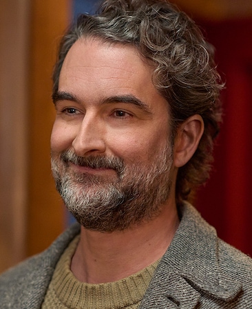 Headshot of Jay Duplass as Steve wearing a grey coat with a orange background.